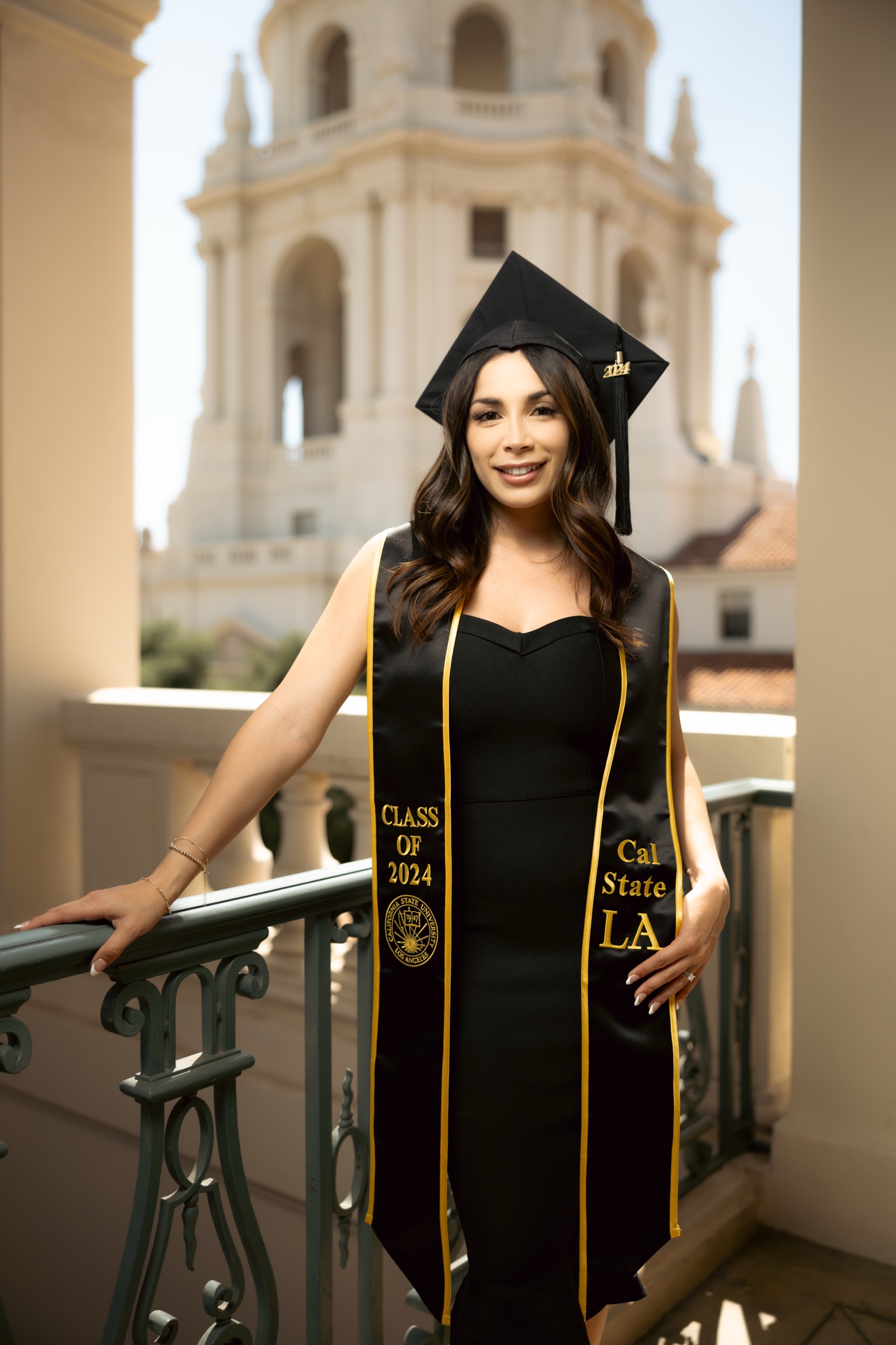 Cal State LA graduate wearing cap and gown on balcony with city tower behind