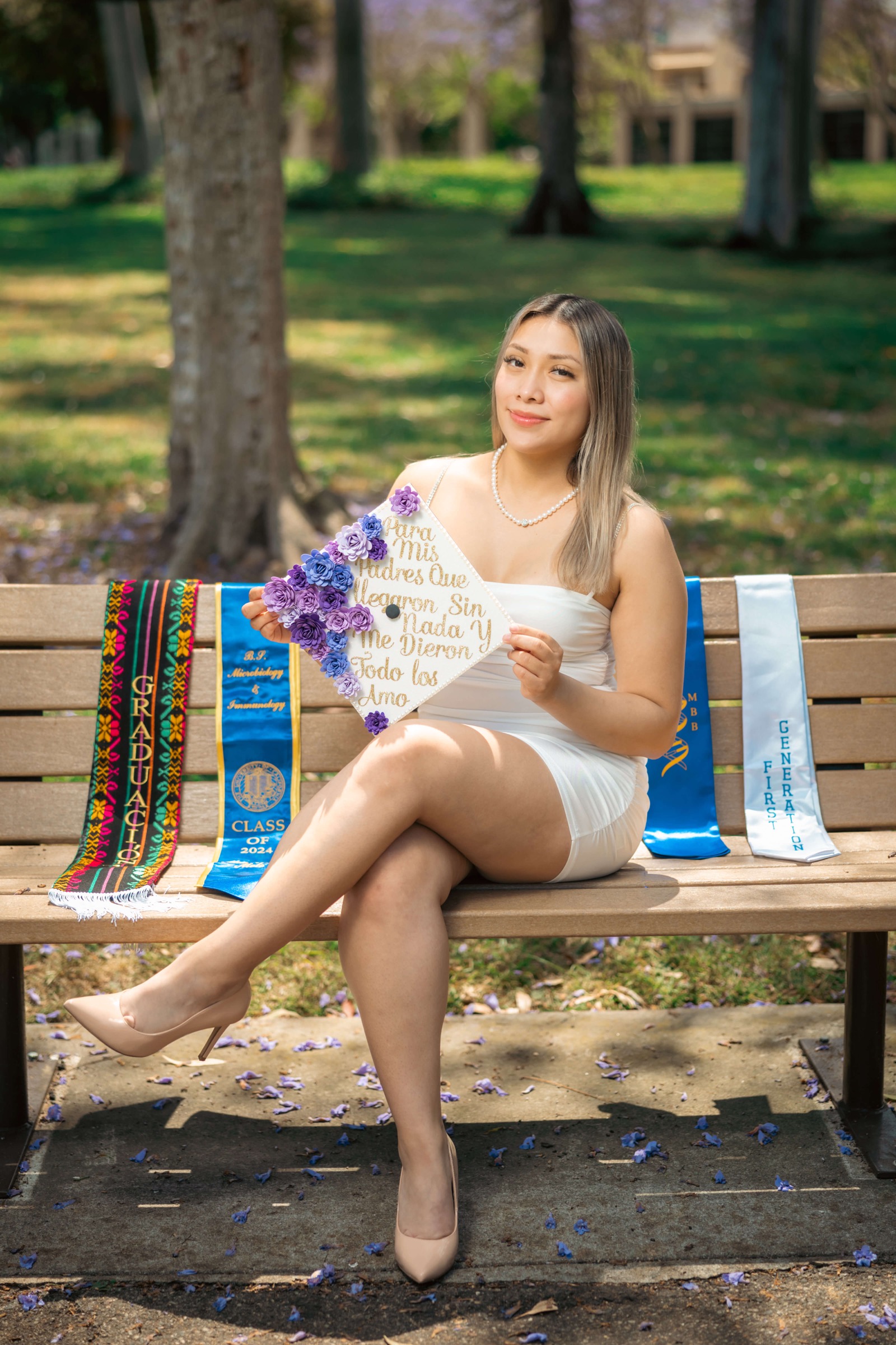 Graduate on park bench with decorated cap, stoles, and cultural sashes laid out