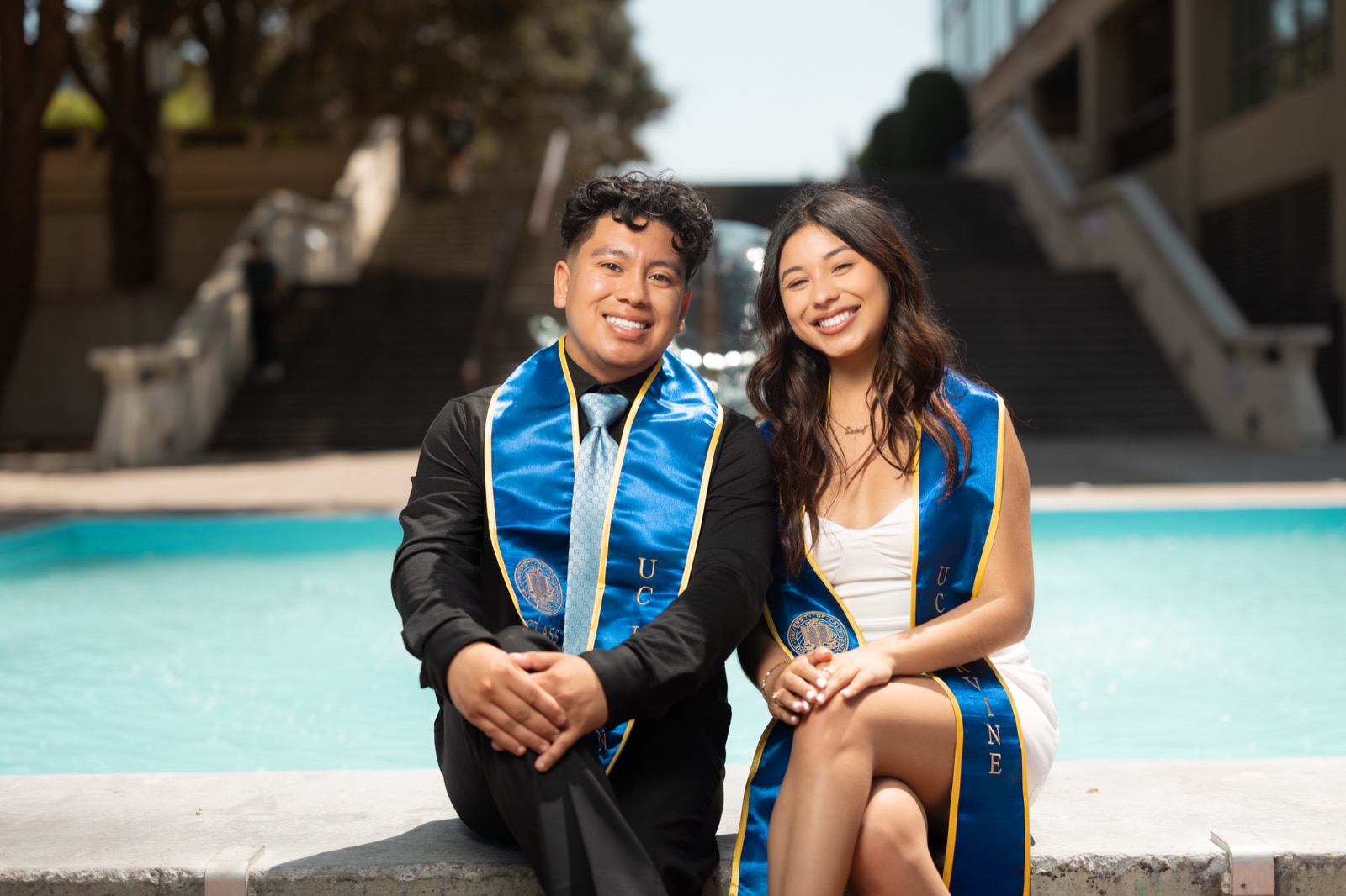 UCI couple sitting together by campus fountain with matching sashes