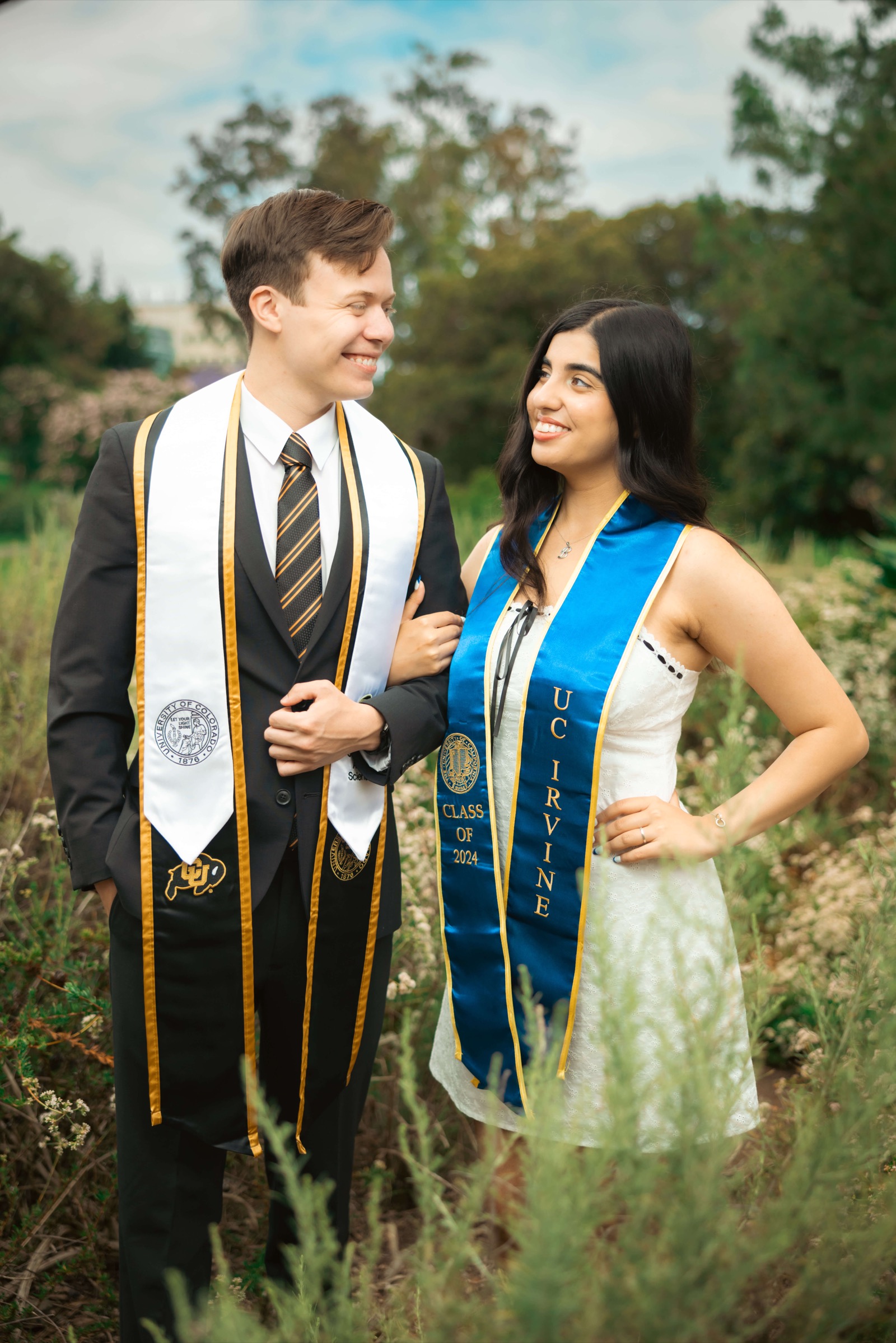 UCI couple in graduation attire smiling at each other in wildflower field