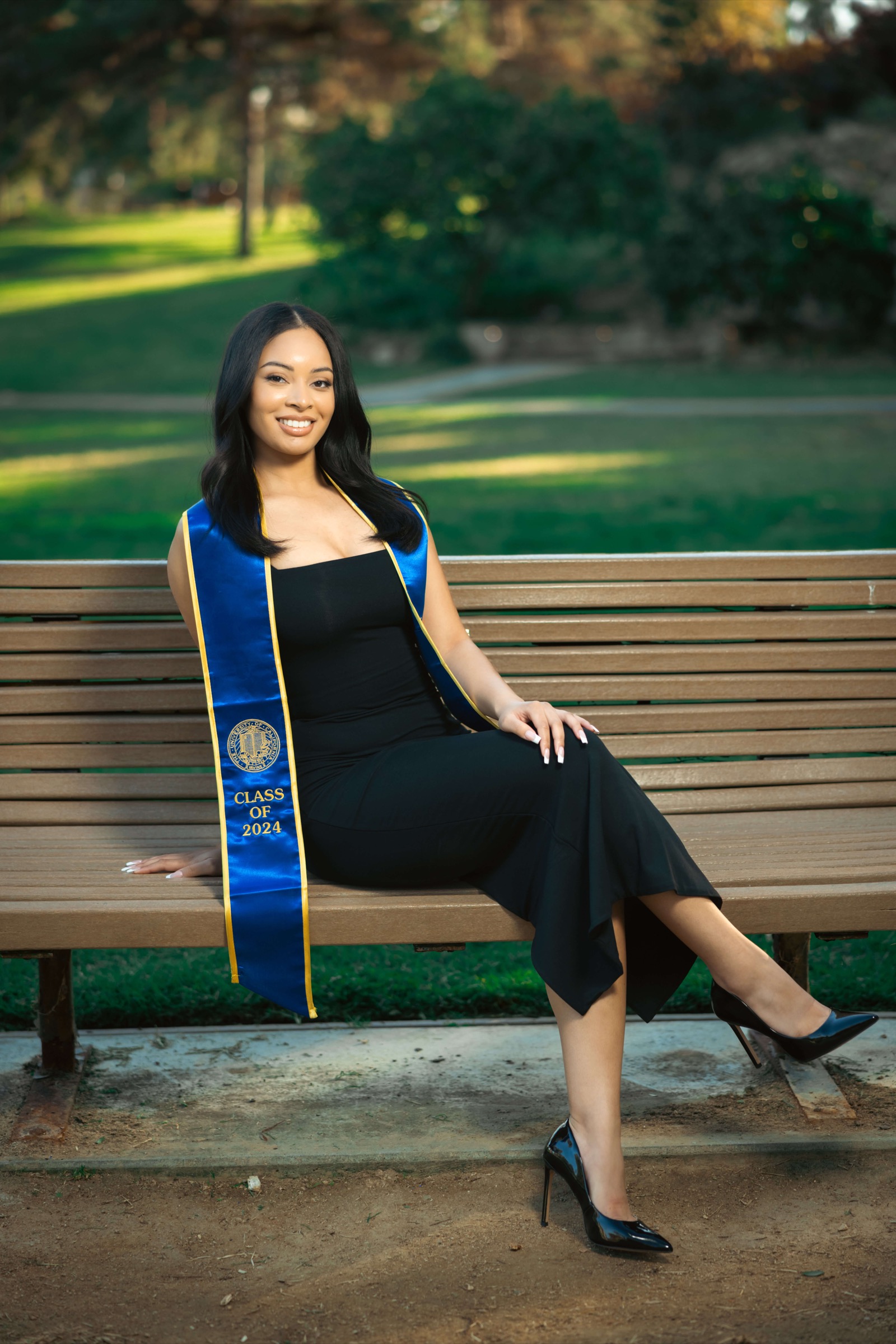 UCI graduate in black dress sitting on park bench with sash